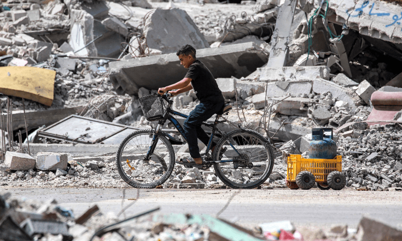 A boy rides a cycle pulling a cart loaded with a cooking gas canister past the rubble of a collapsed building in Khan Younis in the southern Gaza Strip on Sept 16, 2024. — AFP