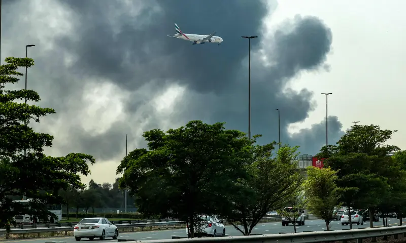 An Emirates aircraft prepares for landing as a smoke plume rises from an ongoing fire near Dubai International Airport in Dubai on March 16, 2026. &mdash; AFP