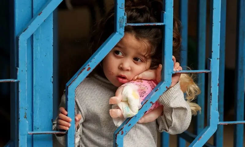 A girl, displaced from southern Lebanon amid the conflict with Israel, holds a doll as she looks through a fence at a school-turned shelter, near Sidon, Lebanon, May 17, 2026. &mdash;AFP
