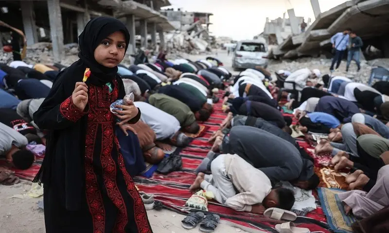 A girl holding a piece of candy stands among Palestinians performing morning prayers marking the start of Eidul Azha, on a heavily damaged street in Khan Yunis in the southern Gaza Strip on May 27, 2026.