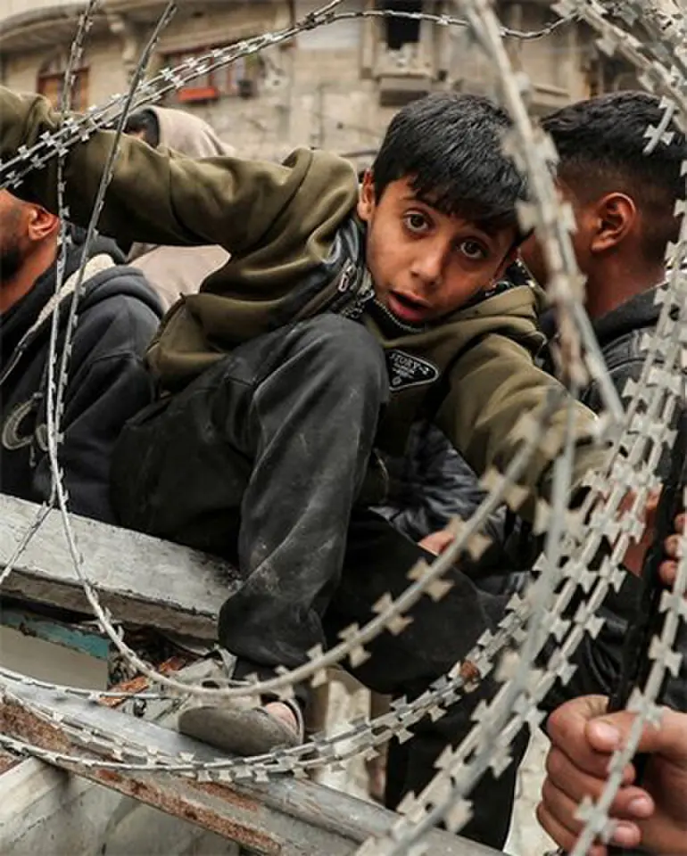 A boy looks through coils of barbed wire as he waits in a line to buy bread in Gaza city, in early 2025, amid the ceasefire between Hamas and Israel | Reuters