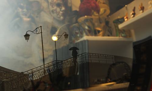 A man, reflected in a carnival masks shop display window, crosses a bridge at the Murano island in Venice on March 2. &mdash; AP