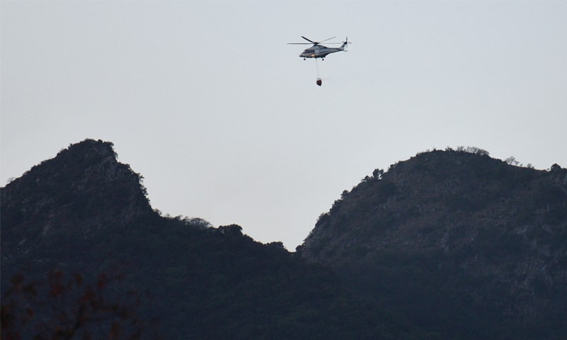 A helicopter, carrying a bucket filled with water, does sorties to extinguish the fire in the Margalla Hills on Friday. — Photo by Tanveer Shahzad