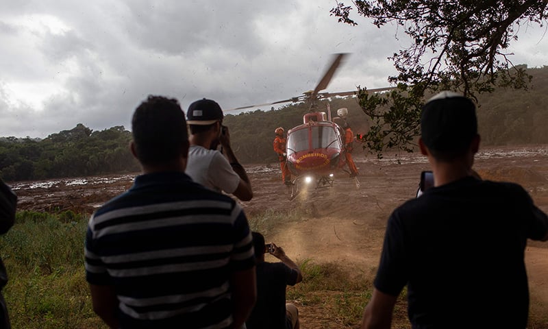 Local residents of the community of Parque das Cachoeiras watch a rescue mission helicopter take off from the mud-covered area a day after the collapse of a dam at an iron-ore mine belonging to Brazil's giant mining company Vale. ─ AFP