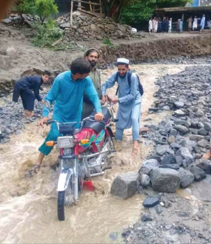 A motorcyclist is helped by two men to cross a portion of a road swept away by flash flood in Gawaldi union council of Upper Dir on Thursday. — Dawn