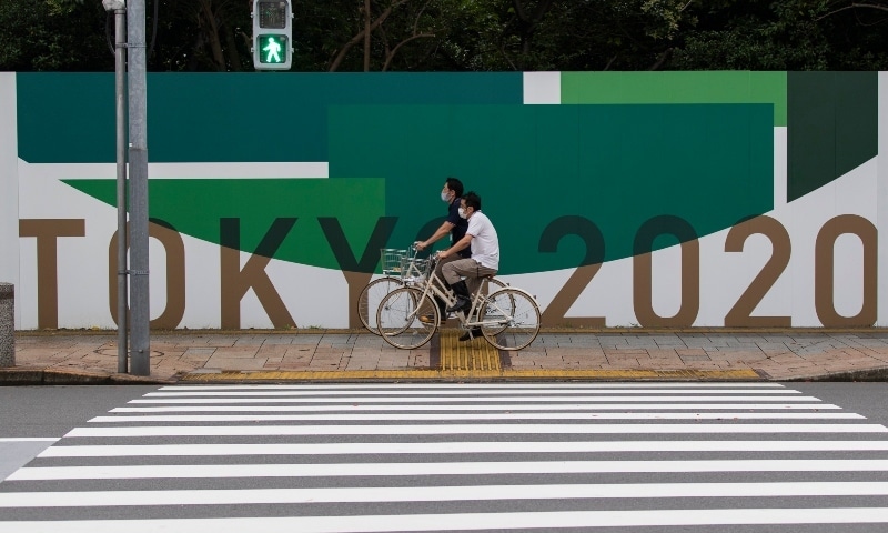 Men wearing face masks ride bicycles along the wall installed to close off a park being prepared for the Olympics and Paralympics Games in Tokyo on July 1. — AP