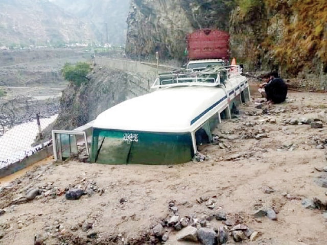 Men sit beside a coach buried under a landslide on KKH in Khushi area of Upper Kohistan on Wednesday. — Dawn