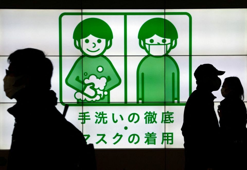 Passersby wearing protective face masks walk past in front of an electric screen displaying notice about Covid-19 safety measures in Tokyo, Japan, February 1. &mdash; Reuters