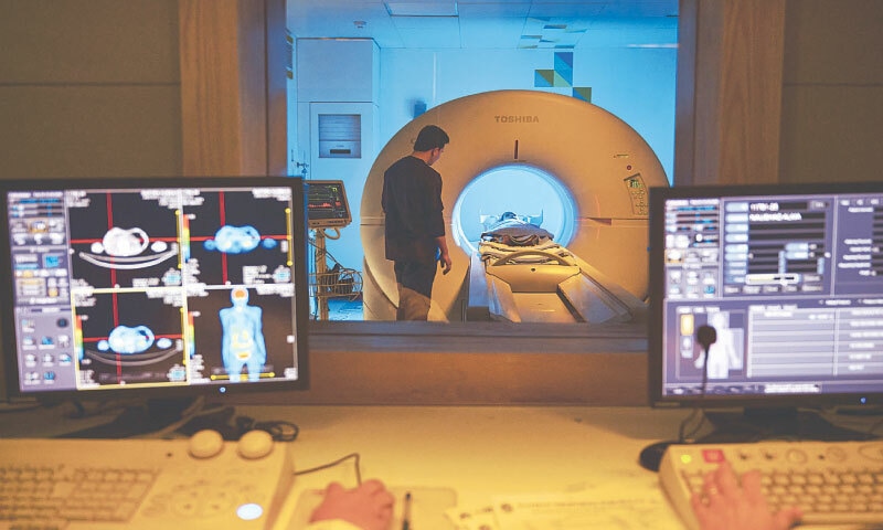 A radiology technologist stands over a patient undergoing a PET CT scan, with results visible on the monitor in adjoining room | Patients’ Aid Foundation