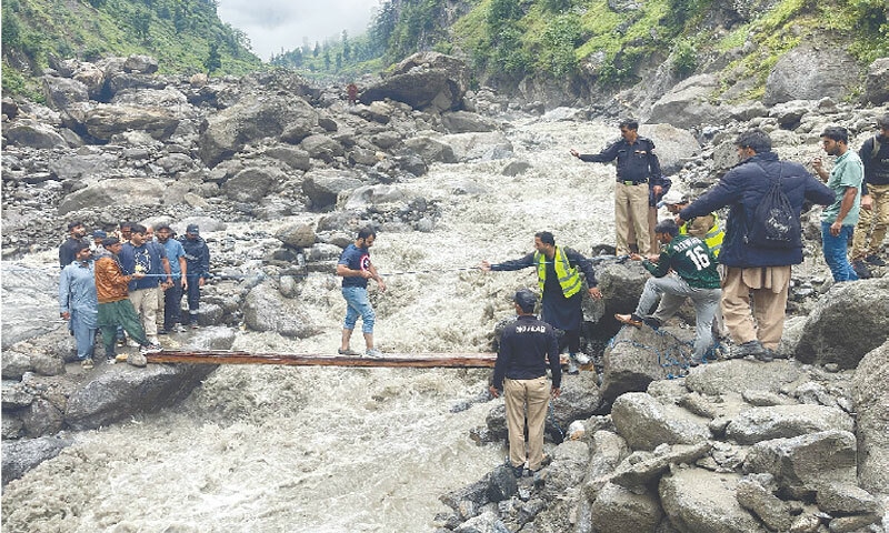 Rescue workers and police personnel help tourists cross a raging torrent on an improvised wooden plank during the evacuation from Ratti Galli lake base camp.&mdash;Dawn