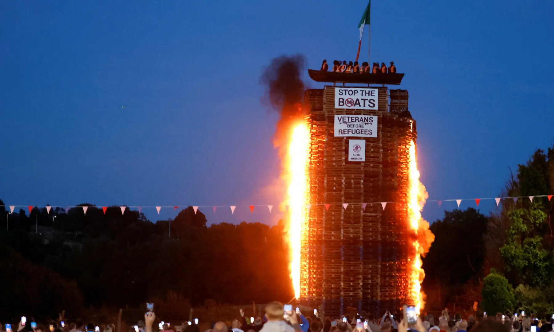 A loyalist bonfire burns after being set alight, featuring a model effigy of a boat containing mannequins portrayed as migrants in lifejackets, with a sign reading &ldquo;Stop the Boats&rdquo;, during annual bonfire events marking the 1690 Battle of the Boyne, in Moygashel, Northern Ireland on July 10, 2025. &mdash; Reuters/Clodagh Kilcoyne/File