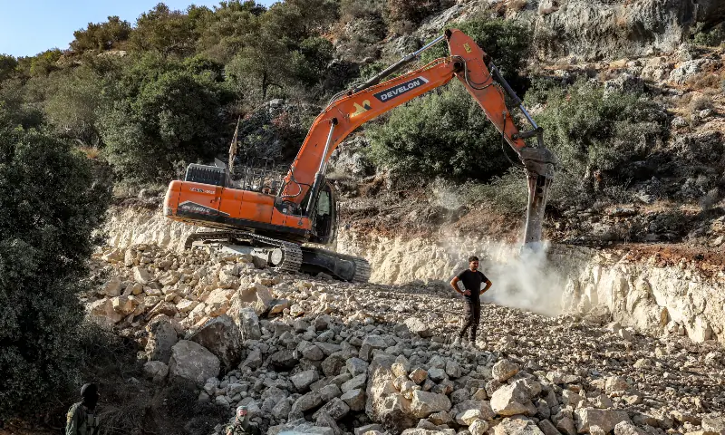 Israeli army soldiers stand guard while an Israeli settlers stands before an excavator clearing soil for a new road under construction for the use of Israeli settlers near the Palestinian village of Beit Ur al-Fauqa, west of Ramallah in the occupied West Bank, on September 29, 2025.  — AFP