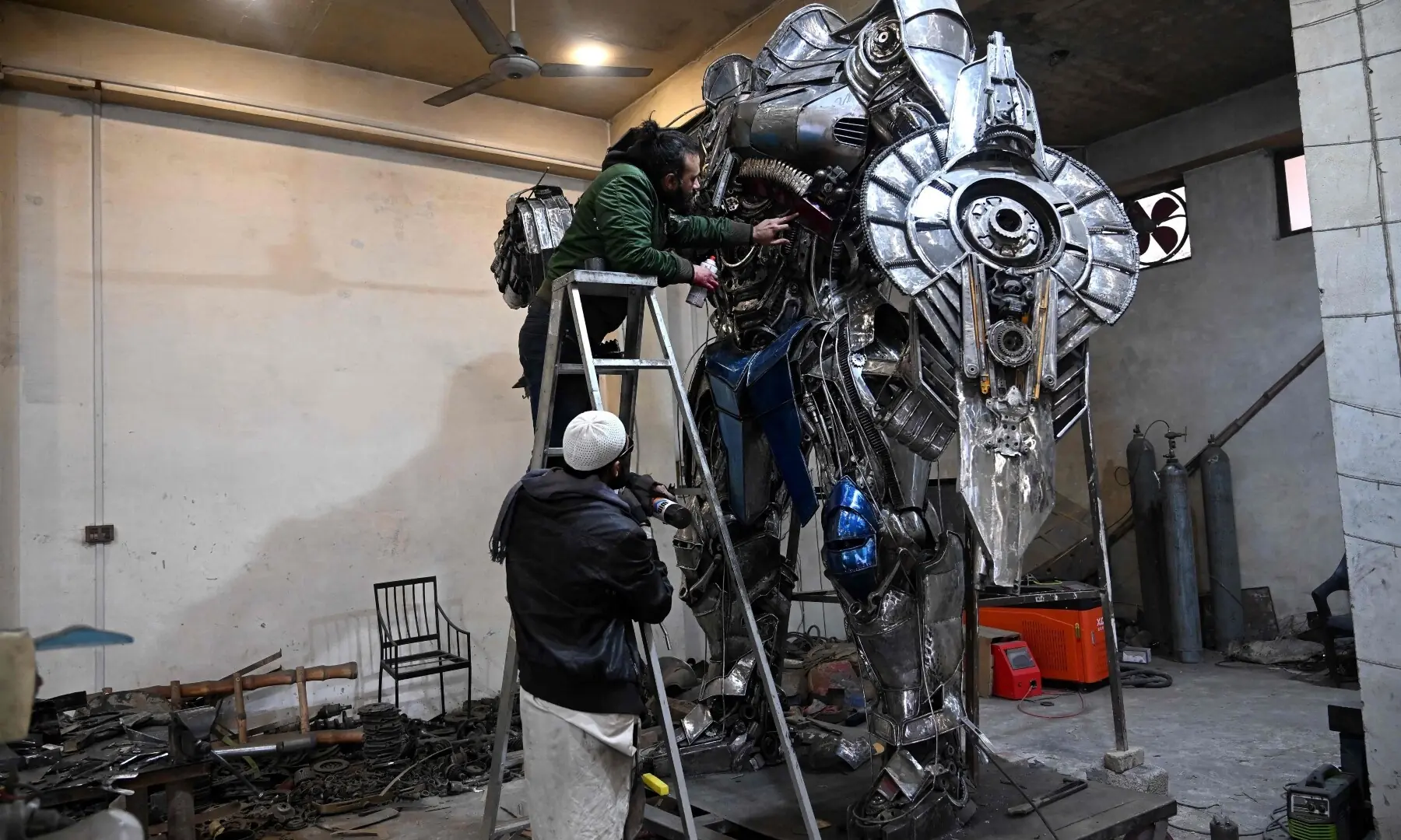 This photograph taken on January 30, 2026 shows sculptor Ehtisham Jadoon (top) spray painting a model of Optimus Prime, a film character from Transformers, made from scrap metal at his studio on the outskirts of Islamabad. &mdash; AFP