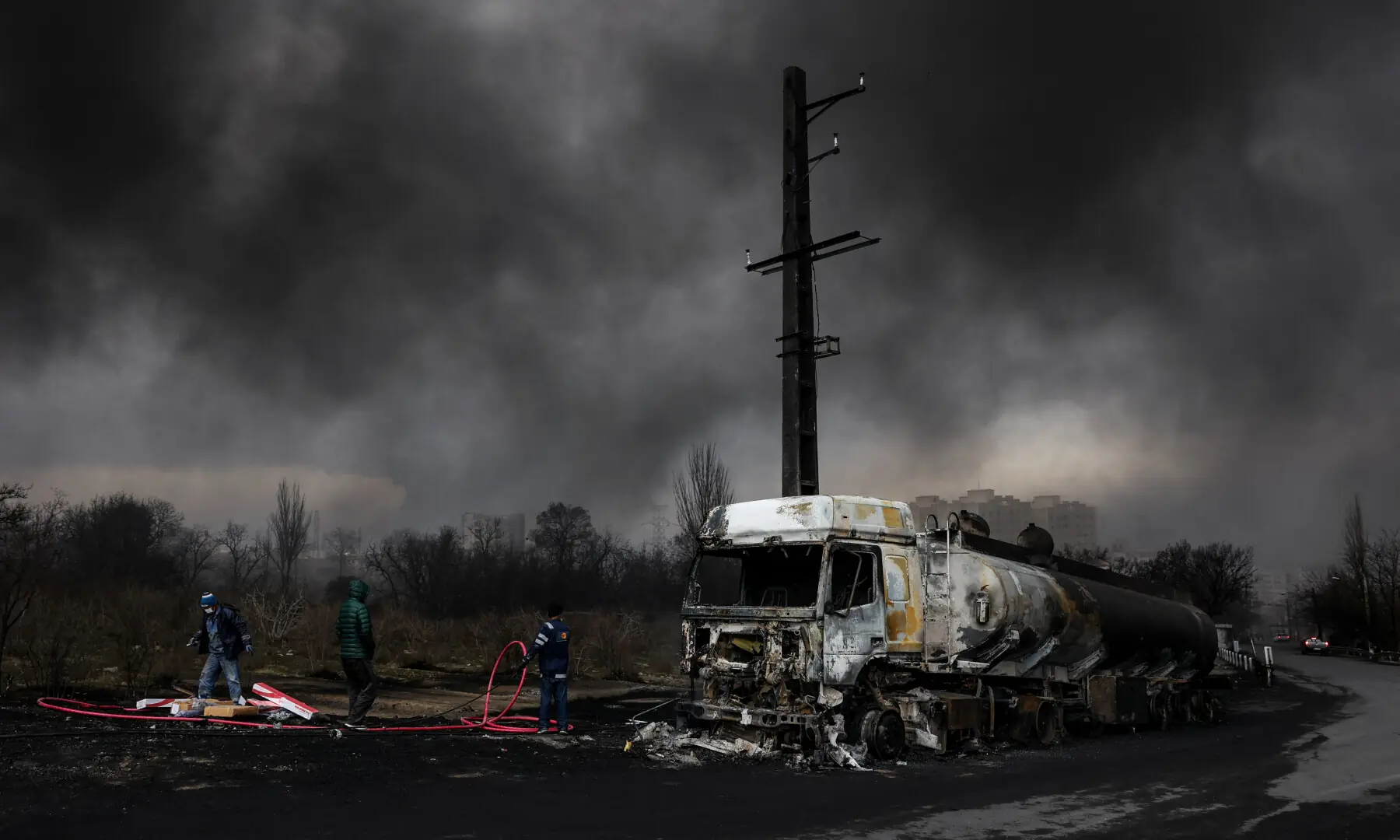 People stand near a destroyed vehicle as smoke rises after a reported strike on Shahran fuel tanks, on March 8, 2026. &mdash; WANA via Reuters