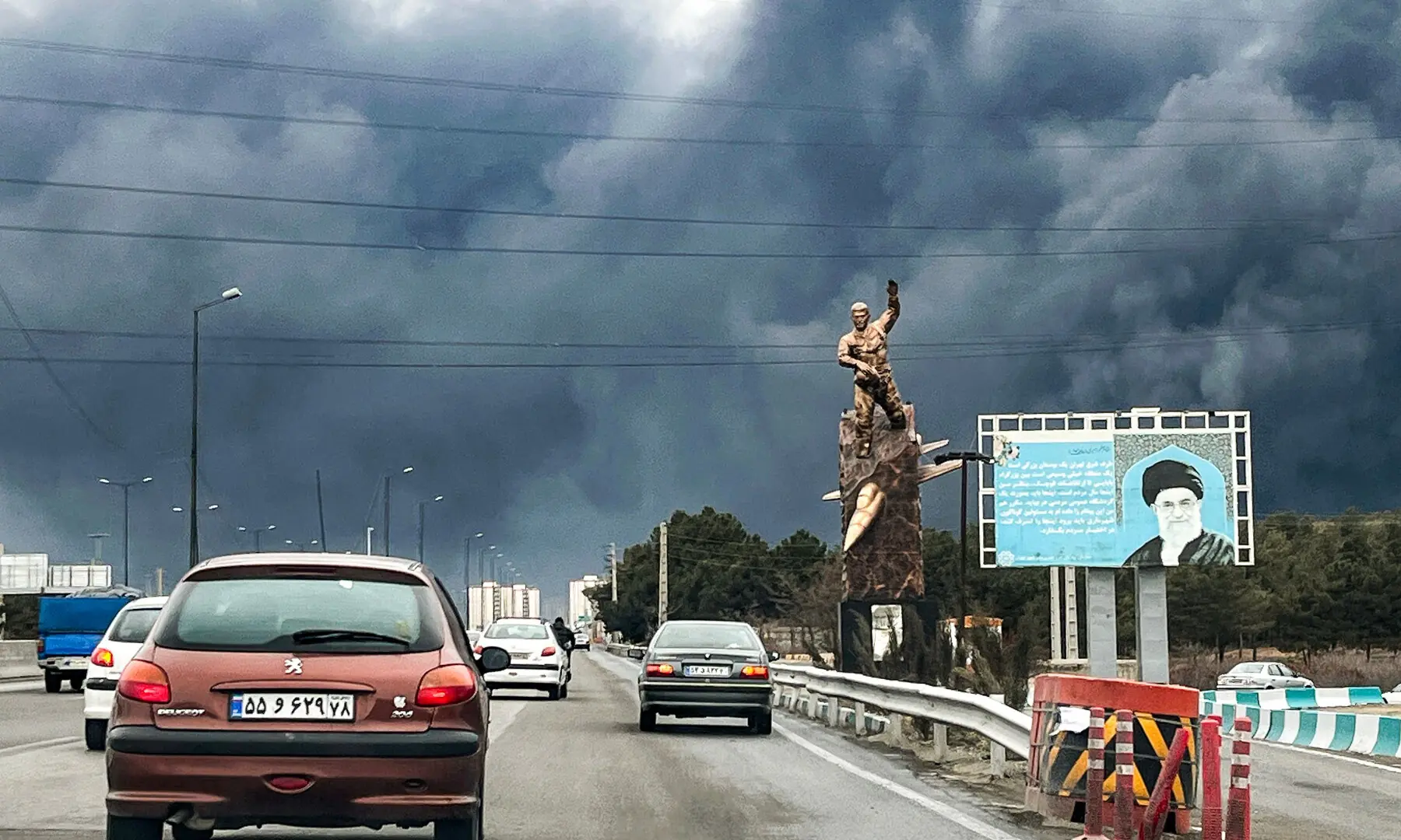 Vehicles move along a highway past a war memorial statue and a billboard depicting Iran&rsquo;s late supreme leader Ayatollah Ali Khamenei, with plumes of black smoke billowing, in Tehran on March 8, 2026. &mdash; AFP