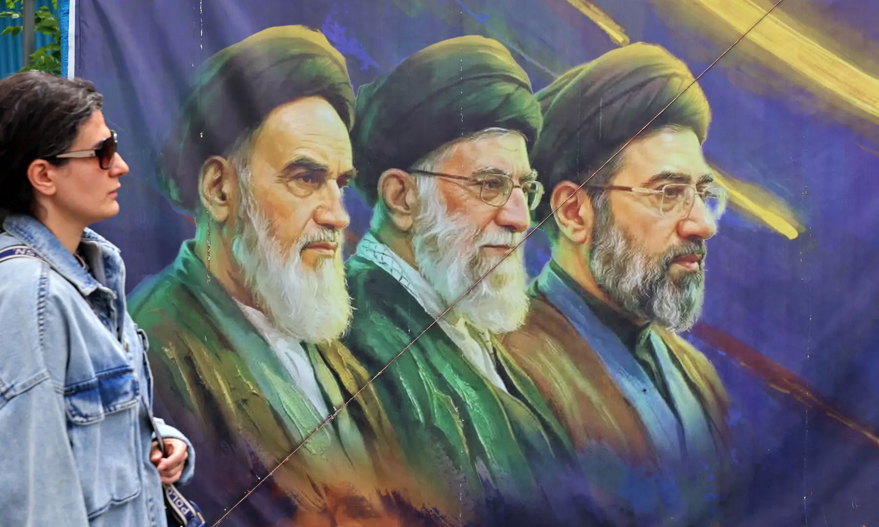 This photo taken on April 19, 2026 shows a woman walking past a banner featuring Iran&rsquo;s late supreme leaders Ayatollah Ruhollah Khomeini (L) and Ayatollah Ali Khamenei (C) next to newly elected supreme leader Ayatollah Mojtaba Khamenei, in Tehran.  &mdash; AFP/File