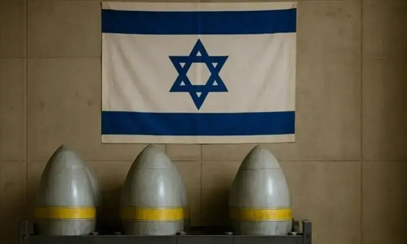 Image depicting a symbolic military setting with the flag of Israel hanging on a neutral-colored, tiled wall. Below the flag are three large military projectiles (missiles or artillery shells) standing upright in a rack. &mdash; Anadolu Agency/File