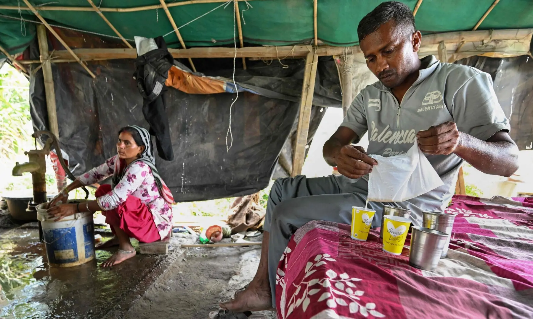 This photograph taken on June 3, 2026 shows Debvati (L) washing clothes as her husband farmer Bhole Shankar pours fruit juice after returning from work in approximately 40 degrees Celsius temperatures, inside their shanty on the Yamuna river floodplains in New Delhi. — AFP