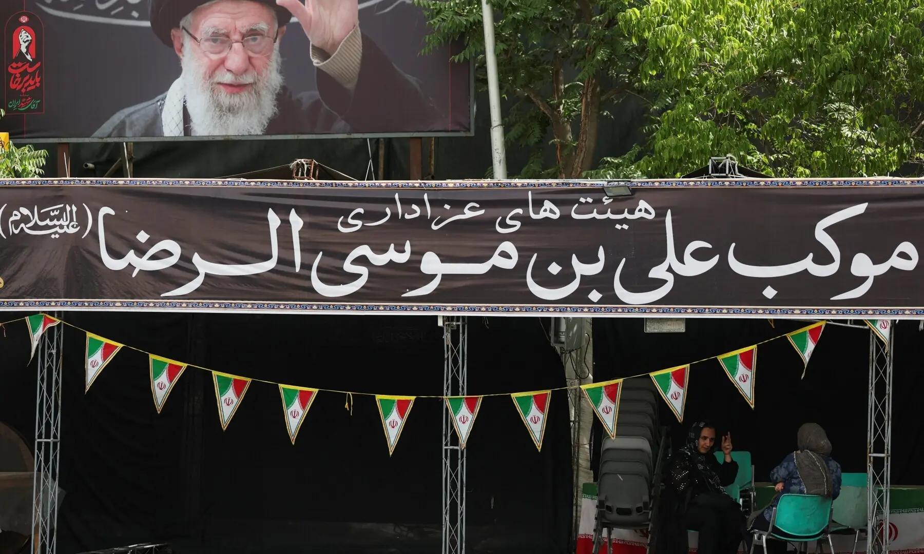 Women sit under a poster with the image of Iran&rsquo;s assassinated Supreme Leader Ayatollah Ali Khamenei, in Tehran, Iran on July 2, 2026. &mdash; Reuters
