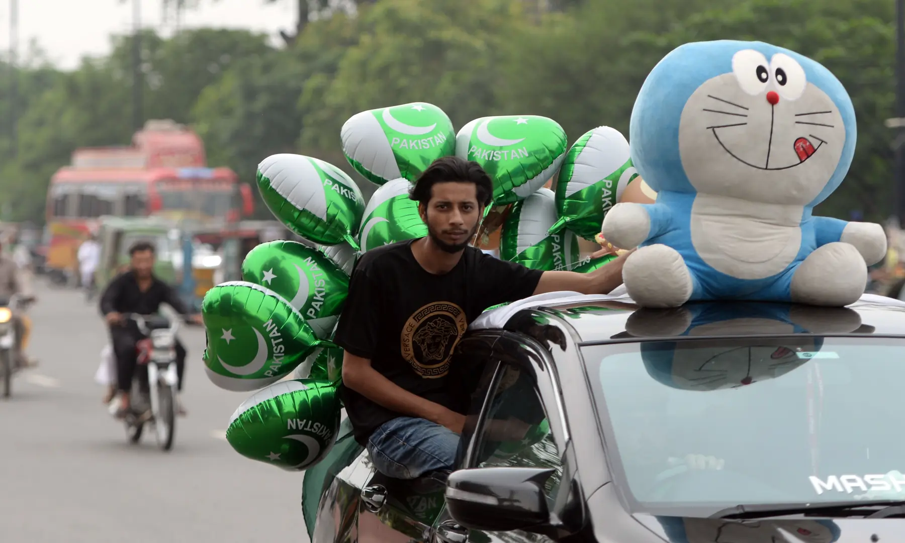 A Doraemon plush and balloons printed with the Pakistan flag are seen tied to a car, while a person sits on the edge of the window, driving along Mall Road in Lahore on Aug 14, 2026. — White Star