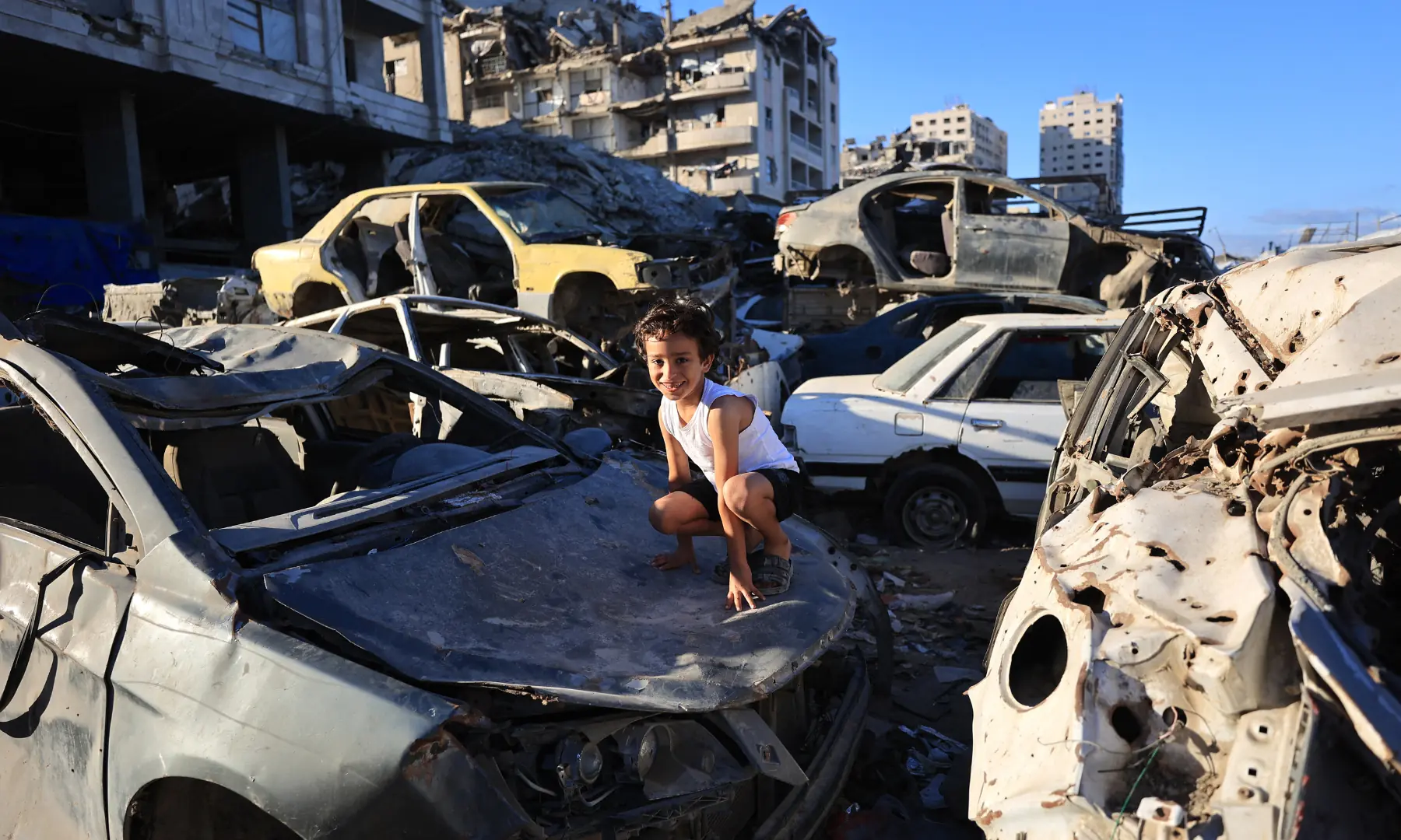 A displaced Palestinian boy plays amid stacked damaged cars against the backdrop of destroyed buildings in Gaza City on Oct 7, 2026. &mdash; AFP